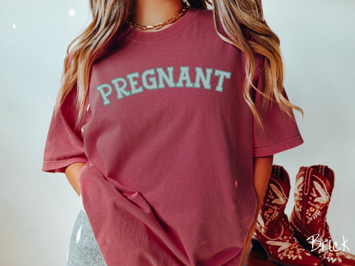A woman wearing a vintage, brick colored Comfort Colors t-shirt with the text PREGNANT in light green and dark green outlined uppercase font.