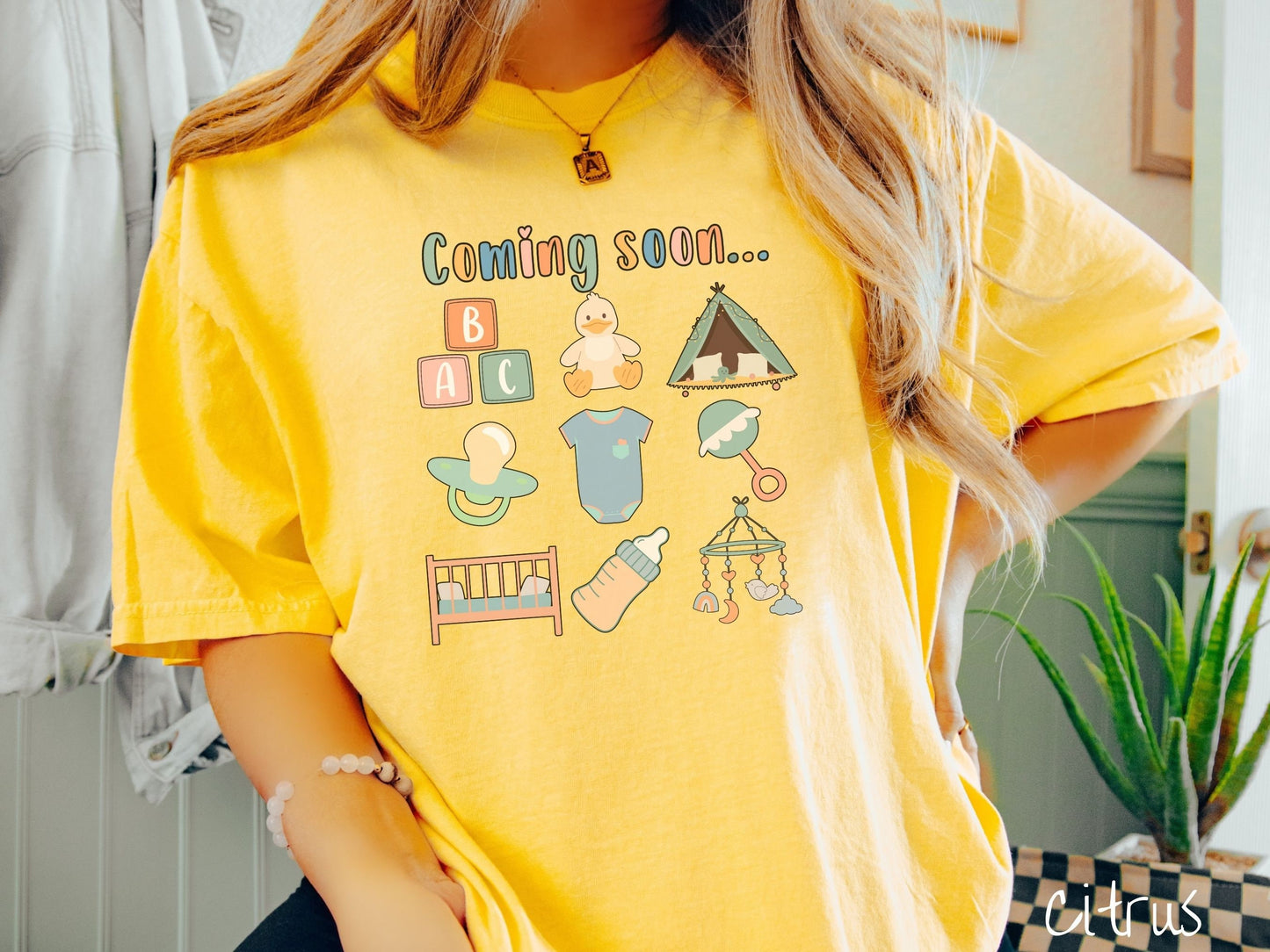 A woman wearing a vintage, citrus colored Comfort Colors t-shirt with the text in colorful font Coming soon… Below this are pictures in a three by three grid of kids toys, binkies, onesies, bottles, a crib, and alphabet blocks.