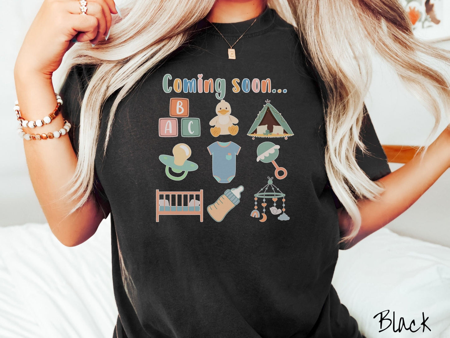 A woman wearing a vintage, black colored Comfort Colors t-shirt with the text in colorful font Coming soon… Below this are pictures in a three by three grid of kids toys, binkies, onesies, bottles, a crib, and alphabet blocks.