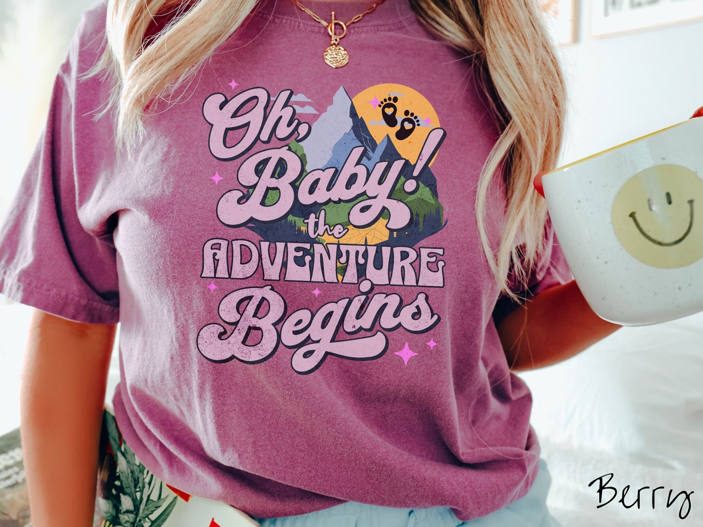 A woman wearing a vintage, berry colored Comfort Colors t-shirt with the text in purple font Oh, Baby! The Adventure Begins! Behind this text are a blue mountain above green meadows, and behind is a yellow moon with black baby footprints inside.