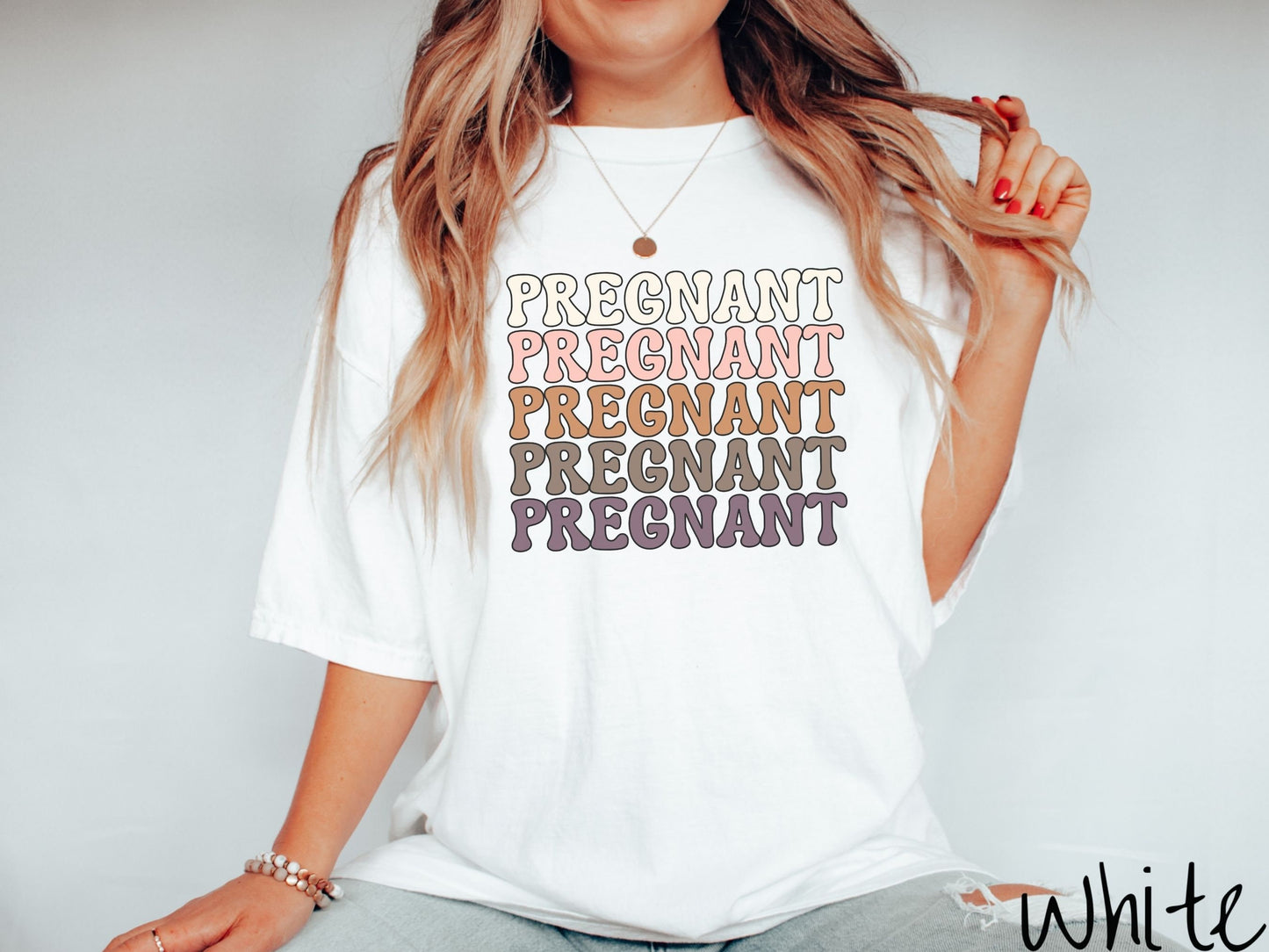 A woman wearing a vintage, white colored Comfort Colors t-shirt with the text PREGNANT in uppercase font listed five times vertically. One is ivory colored, the next pink, next light brown, next dark brown, and the fifth is purple.