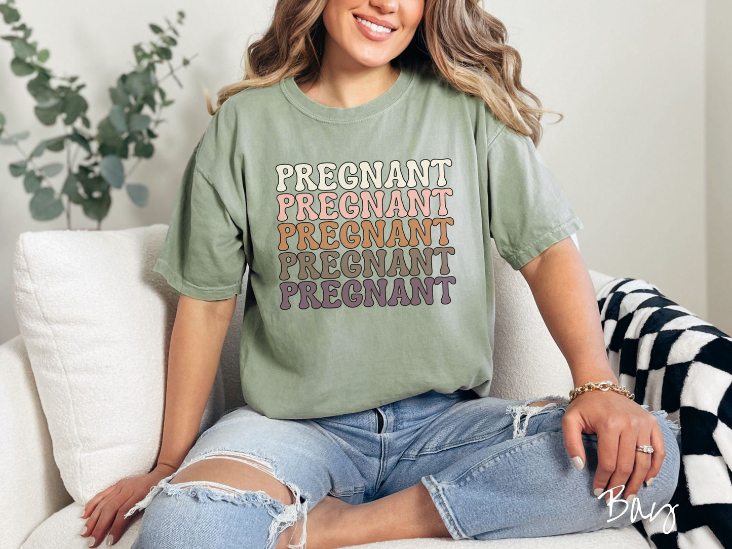 A woman wearing a vintage, bay colored Comfort Colors t-shirt with the text PREGNANT in uppercase font listed five times vertically. One is ivory colored, the next pink, next light brown, next dark brown, and the fifth is purple.