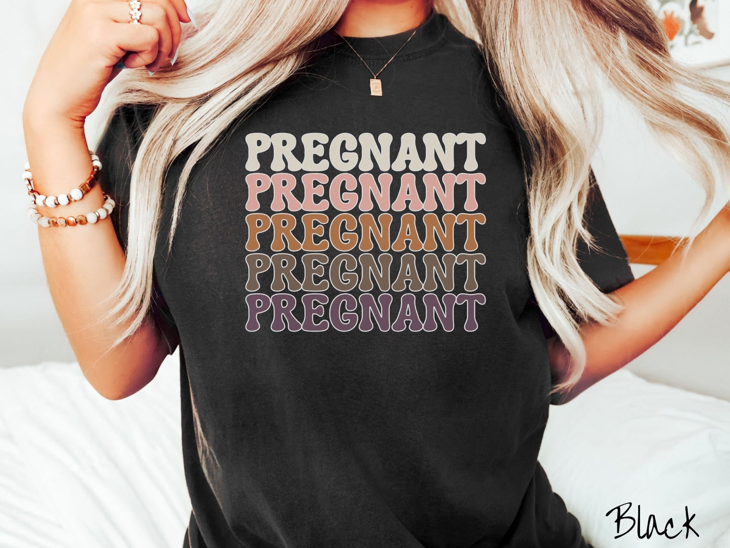 A woman wearing a vintage, black colored Comfort Colors t-shirt with the text PREGNANT in uppercase font listed five times vertically. One is ivory colored, the next pink, next light brown, next dark brown, and the fifth is purple.