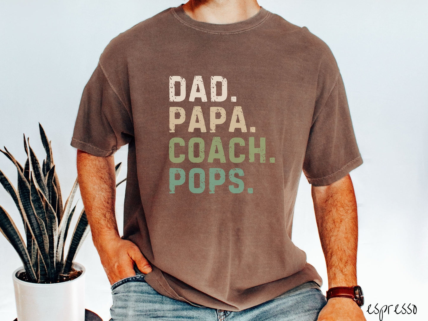 A man wearing a vintage, espresso colored Comfort Colors t-shirt with the text in varying blue colored font Dad. Papa. Coach. Pops.