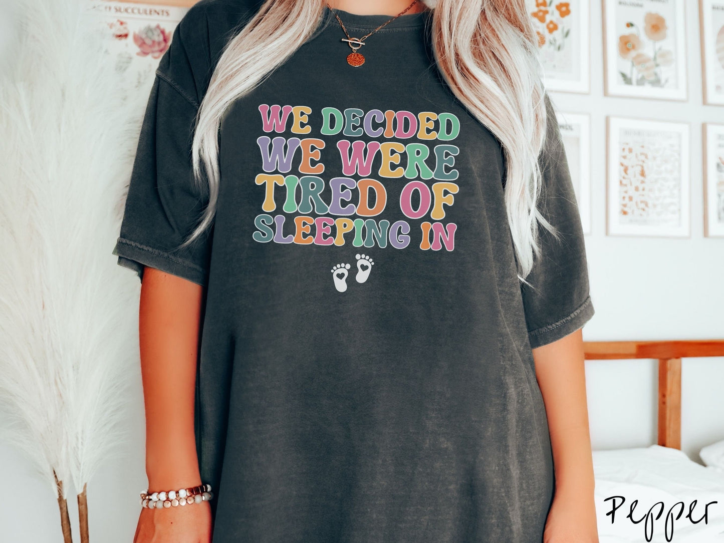 A woman wearing a vintage, pepper colored Comfort Colors t-shirt with the text We Decided We Were Tired Of Sleeping In in pink, yellow, green, and purple uppercase font. Below the text are two white baby footprints with hearts inside them.