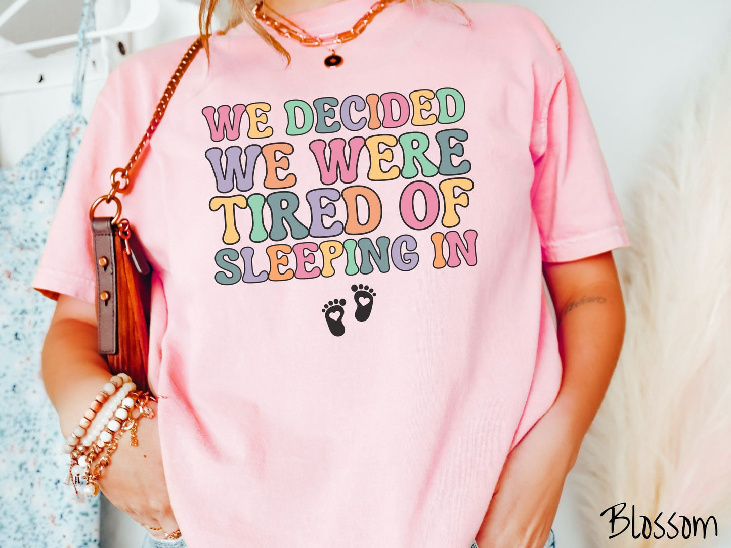 A woman wearing a vintage, blossom colored Comfort Colors t-shirt with the text We Decided We Were Tired Of Sleeping In in pink, yellow, green, and purple uppercase font. Below the text are two white baby footprints with hearts inside them.
