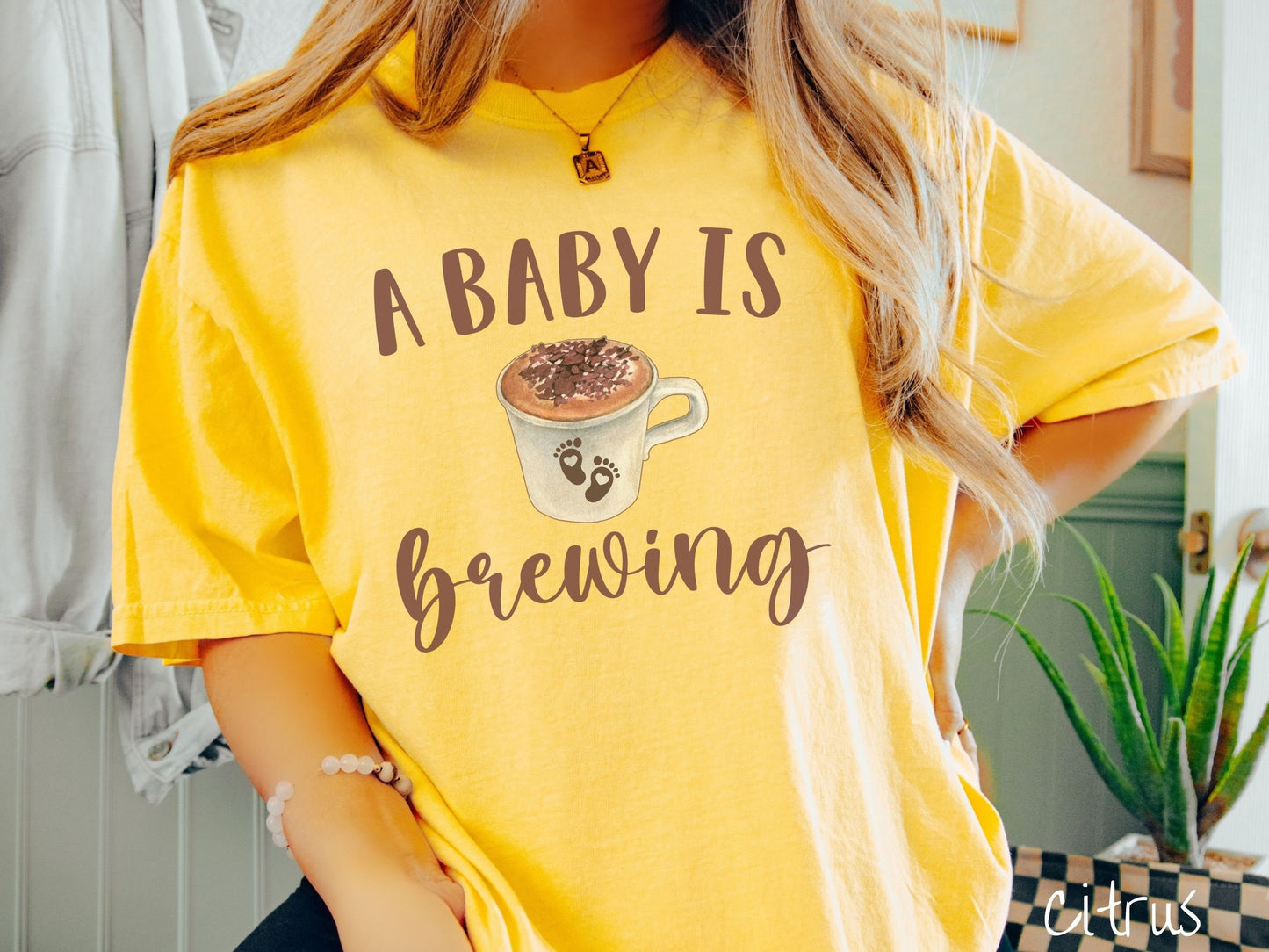 A woman wearing a vintage, citrus colored Comfort Colors t-shirt with the text A Baby Is Brewing in light brown font. In between the text is a white cup of coffee with cream on top. The cup has two baby footprints on it with hearts inside them.