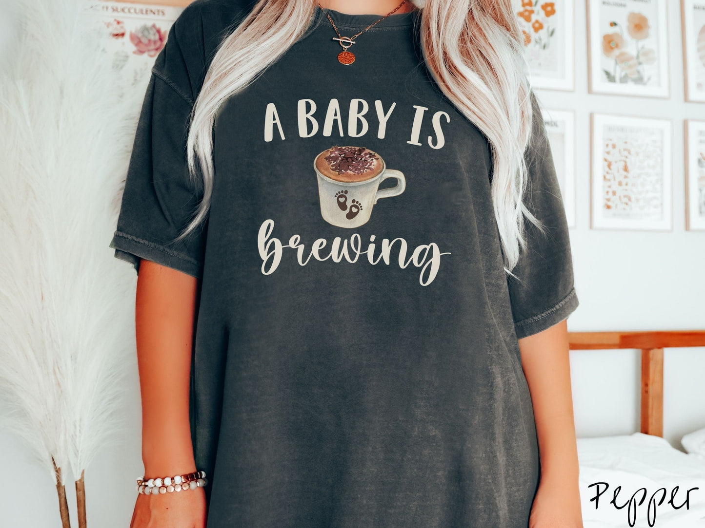 A woman wearing a vintage, pepper colored Comfort Colors t-shirt with the text A Baby Is Brewing in light brown font. In between the text is a white cup of coffee with cream on top. The cup has two baby footprints on it with hearts inside them.
