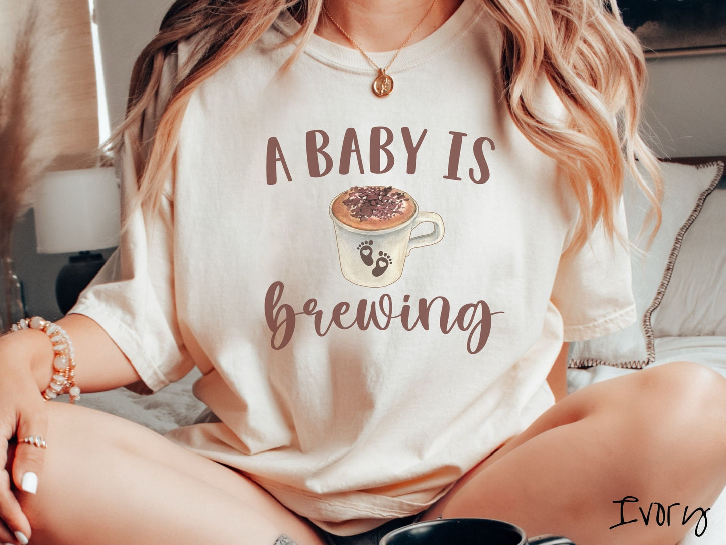 A woman wearing a vintage, ivory colored Comfort Colors t-shirt with the text A Baby Is Brewing in light brown font. In between the text is a white cup of coffee with cream on top. The cup has two baby footprints on it with hearts inside them.