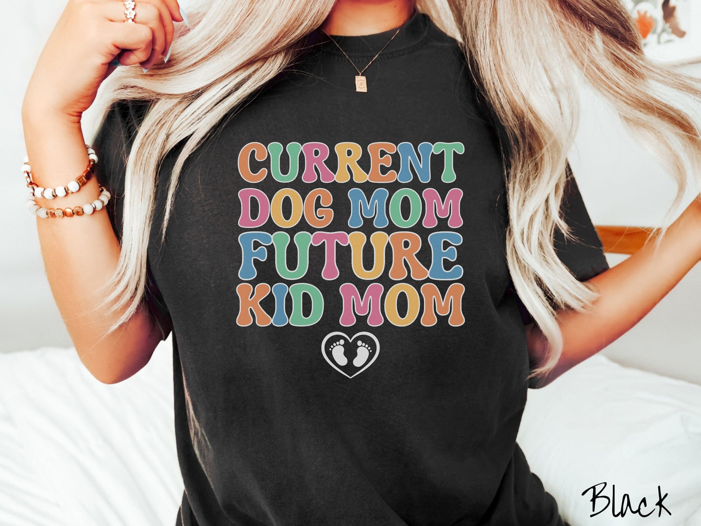 A woman wearing a vintage, black colored Comfort Colors t-shirt with the text Curent Dog Mom Future Kid Mom in orange, green, pink, yellow, and blue uppercase font. Below this is a black outlined heart with two kid footprints inside.