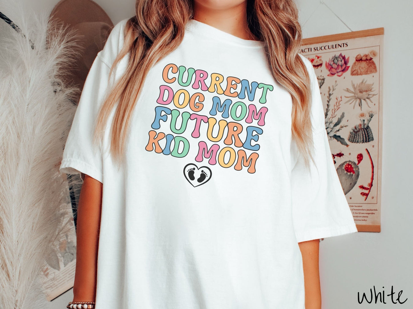 A woman wearing a vintage, white colored Comfort Colors t-shirt with the text Curent Dog Mom Future Kid Mom in orange, green, pink, yellow, and blue uppercase font. Below this is a black outlined heart with two kid footprints inside.