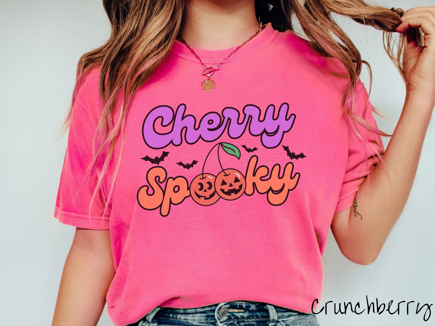 A woman wearing a vintage, crunchberry colored Comfort Colors t-shirt with the text Cherry Spooky in purple and orange font. The two letter O’s in Spooky are smiling jack o'lantern-themed cherries, and there are black bats flying in the background.