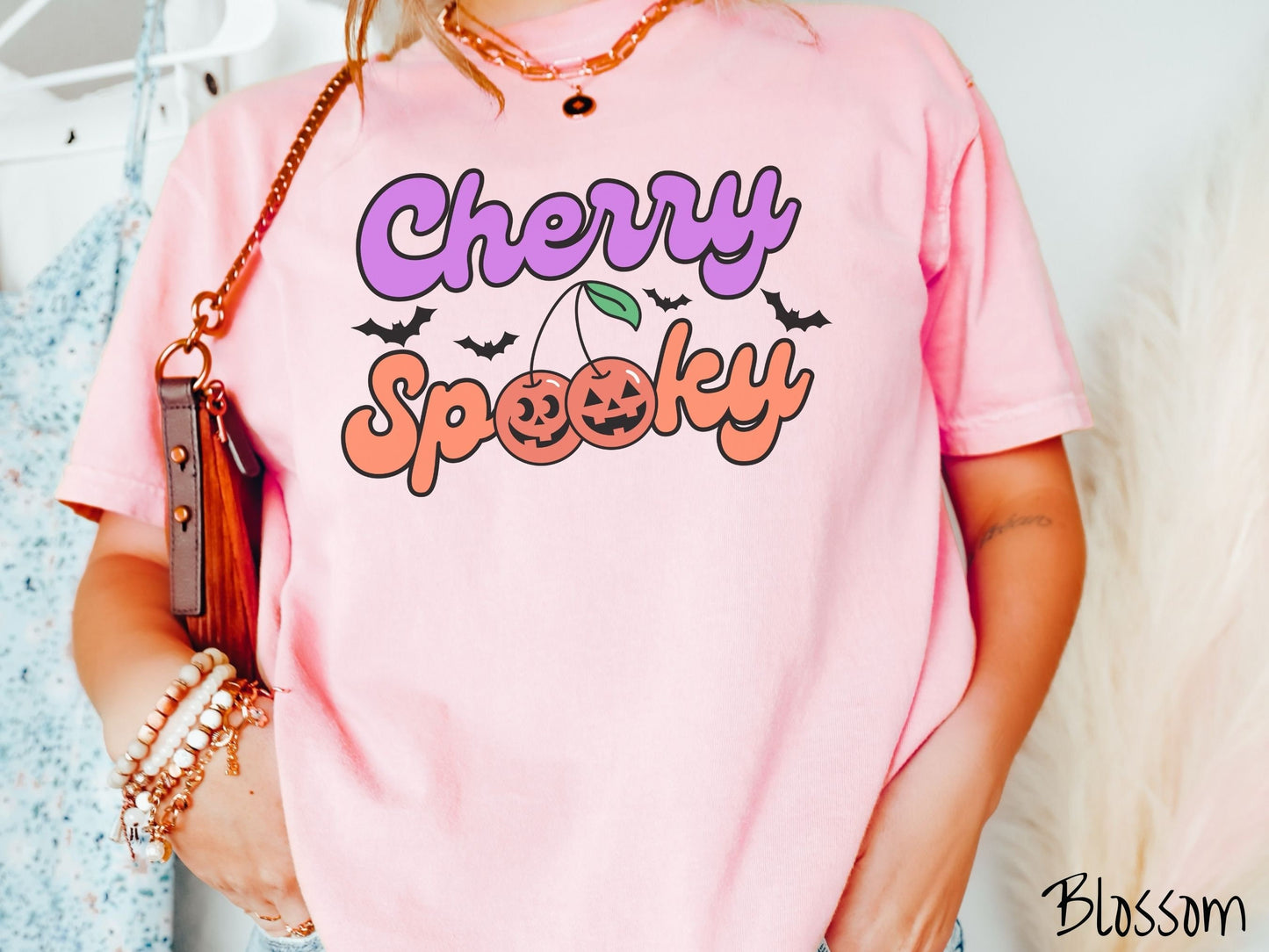 A woman wearing a vintage, blossom colored Comfort Colors t-shirt with the text Cherry Spooky in purple and orange font. The two letter O’s in Spooky are smiling jack o'lantern-themed cherries, and there are black bats flying in the background.