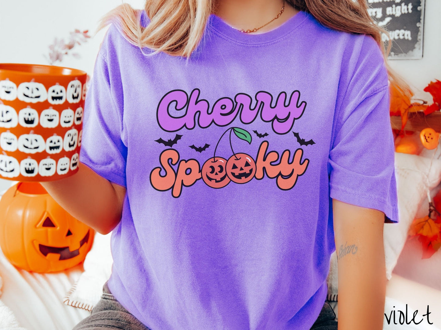 A woman wearing a vintage, violet colored Comfort Colors t-shirt with the text Cherry Spooky in purple and orange font. The two letter O’s in Spooky are smiling jack o'lantern-themed cherries, and there are black bats flying in the background.