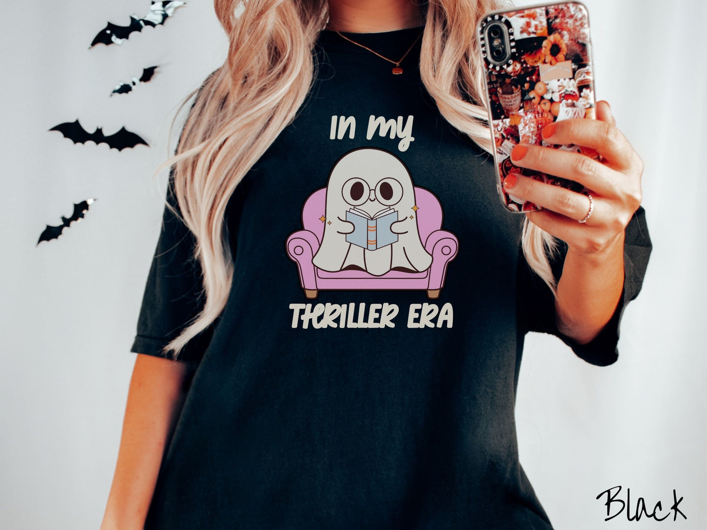 A woman wearing a vintage, black colored Comfort Colors t-shirt with the text In My Thriller Era across the front in ivory font. In between the text is a young ghost with glasses reading a book and sitting in a pink chair.