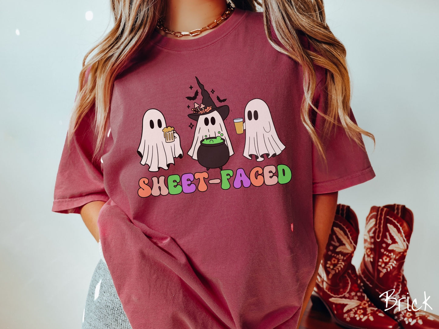 A woman wearing a vintage, brick colored Comfort Colors t-shirt with the text Sheet-Faced in colorful font across the front. Above are three white ghosts drinking beers and standing around a black cauldron with a bubbling green liquid in it.