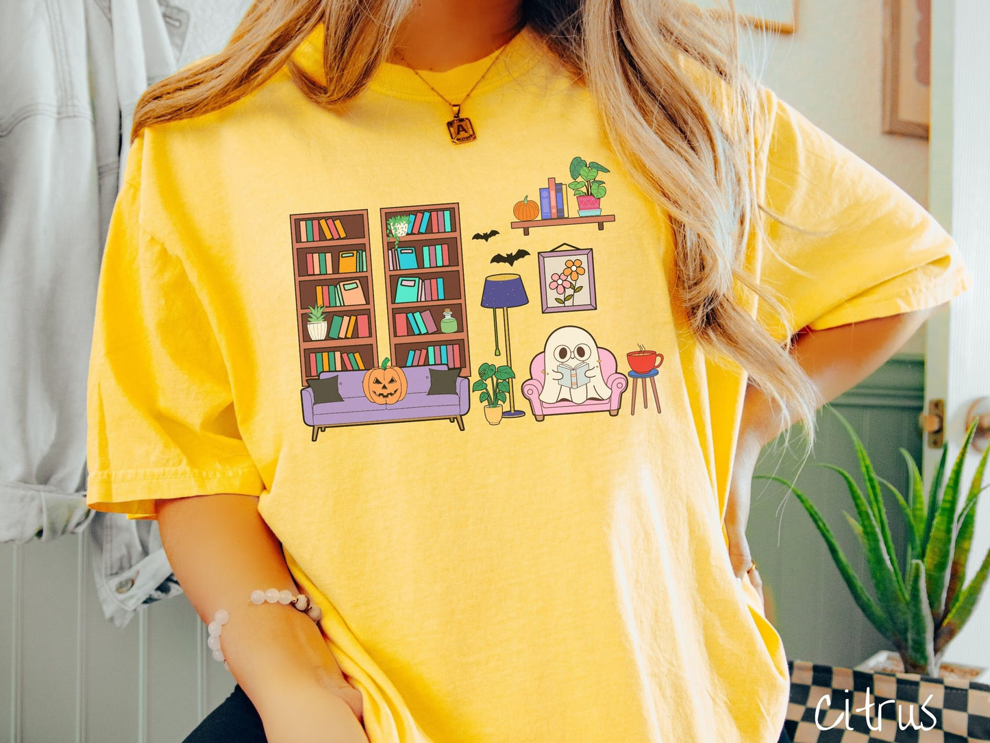 A woman wearing a vintage, citrus colored Comfort Colors t-shirt with a cozy Halloween-themed room. A young white ghost is reading a book in a chair next to a coffee and colorful bookcase.. An orange pumpkin sits on the couch and bats are in the air.