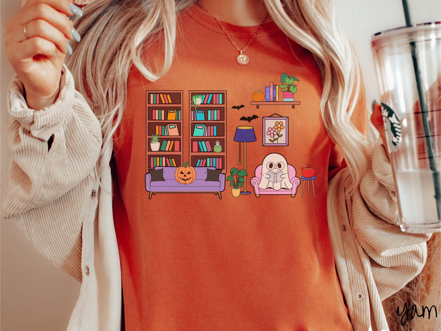 A woman wearing a vintage, yam colored Comfort Colors t-shirt with a cozy Halloween-themed room. A young white ghost is reading a book in a chair next to a coffee and colorful bookcase.. An orange pumpkin sits on the couch and bats are in the air.