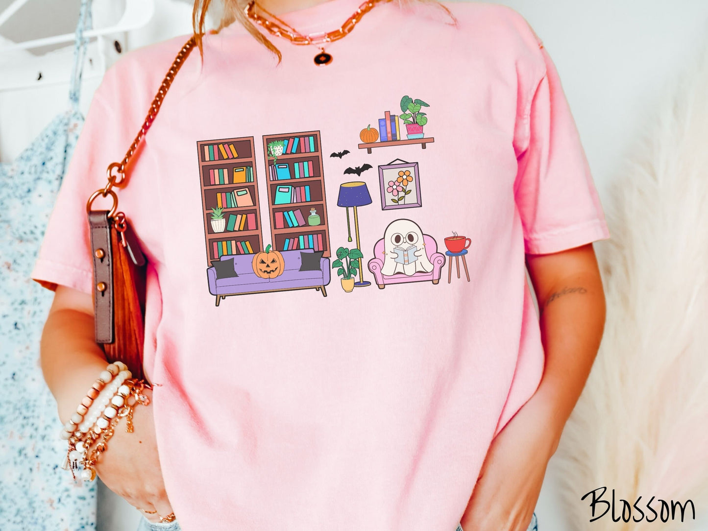 A woman wearing a vintage, blossom colored Comfort Colors t-shirt with a cozy Halloween-themed room. A young white ghost is reading a book in a chair next to a coffee and colorful bookcase. An orange pumpkin sits on the couch and bats are in the air.
