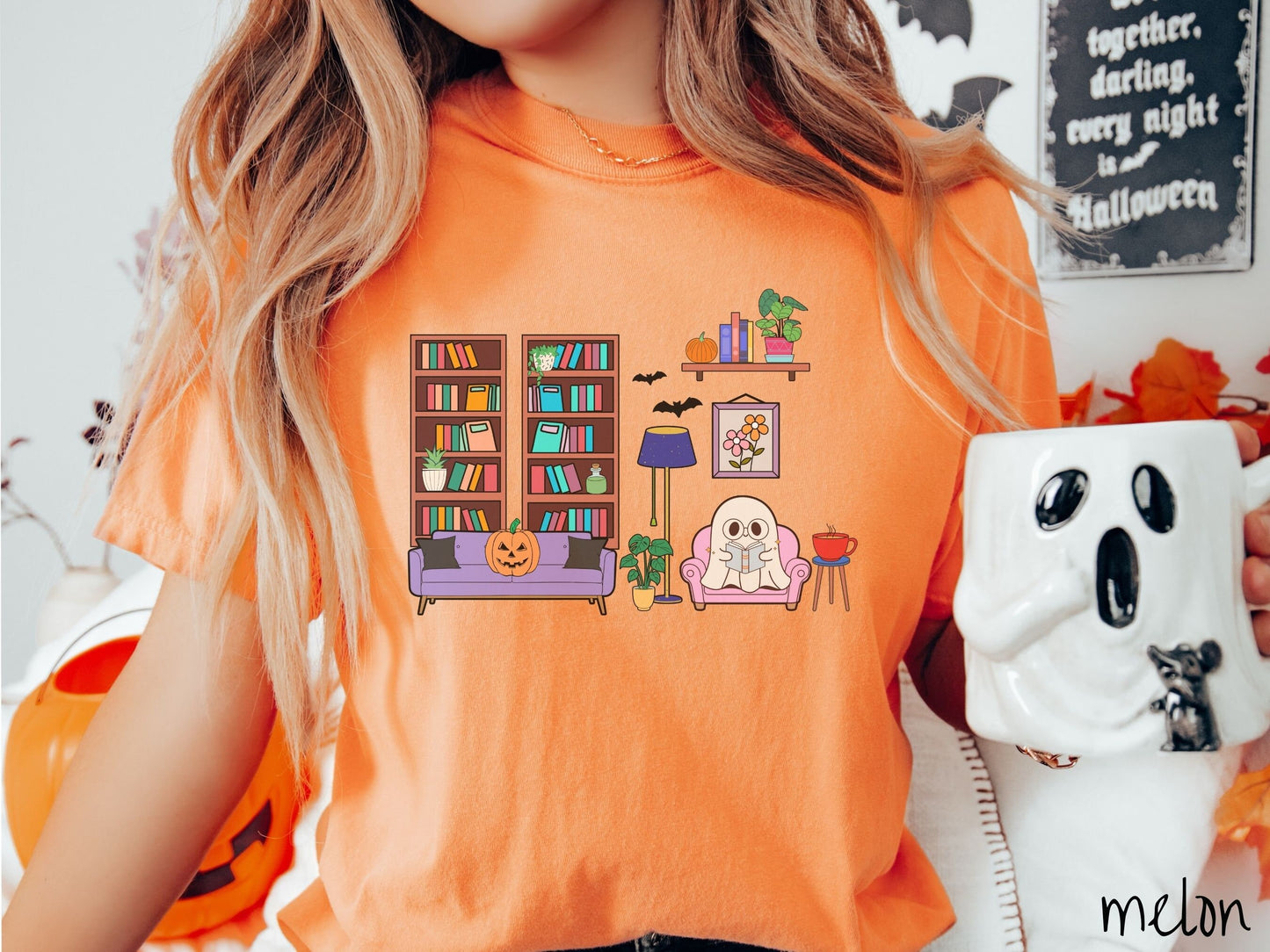 A woman wearing a vintage, melon colored Comfort Colors t-shirt with a cozy Halloween-themed room. A young white ghost is reading a book in a chair next to a coffee and colorful bookcase.. An orange pumpkin sits on the couch and bats are in the air.