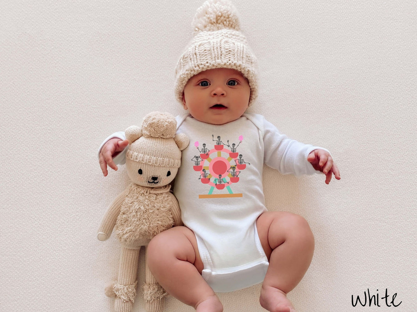 A baby wearing a cute white colored body suit with a ferris wheel filled with black skeletons waving and holding cotton candy as they go around the ride.