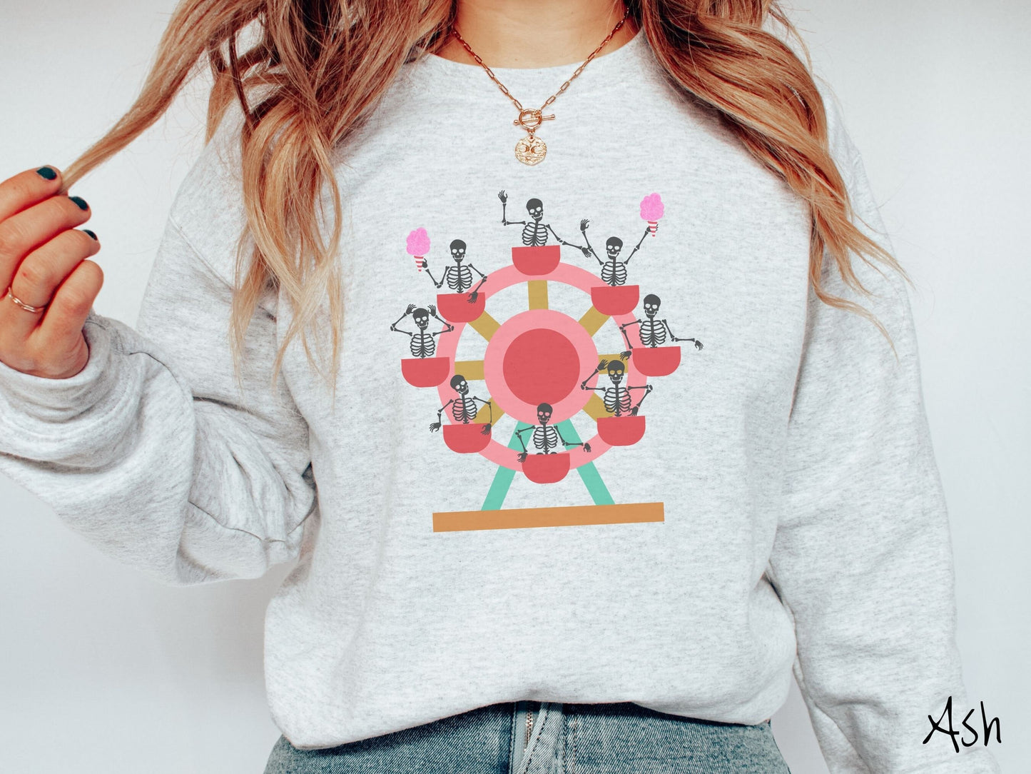 A woman wearing a cute ash colored comfy sweatshirt with a ferris wheel filled with black skeletons waving and holding cotton candy as they go around the ride.