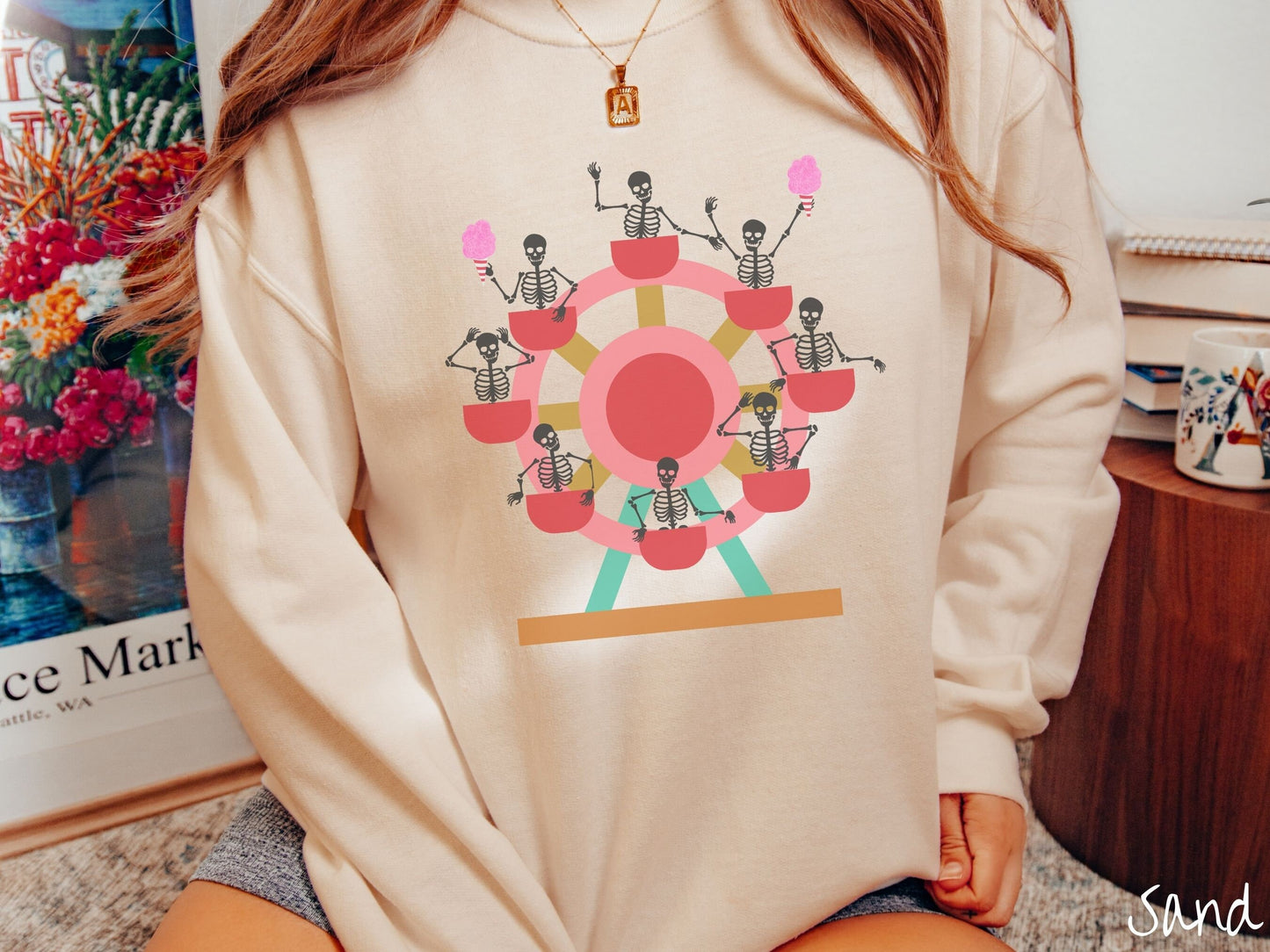 A woman wearing a cute sand colored comfy sweatshirt with a ferris wheel filled with black skeletons waving and holding cotton candy as they go around the ride.