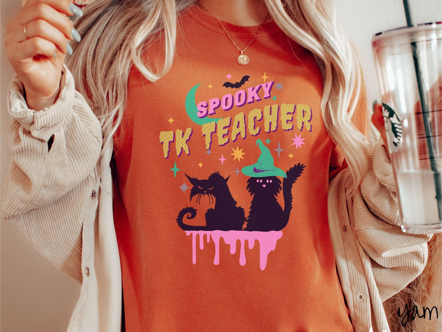 A woman wearing a cute, vintage yam colored Comfort Colors t-shirt with the text Spooky TK Teacher in pink and yellow font. Below are two black cats with pink eyes wearing witch hats sitting beneath colorful stars and a bright green moon.