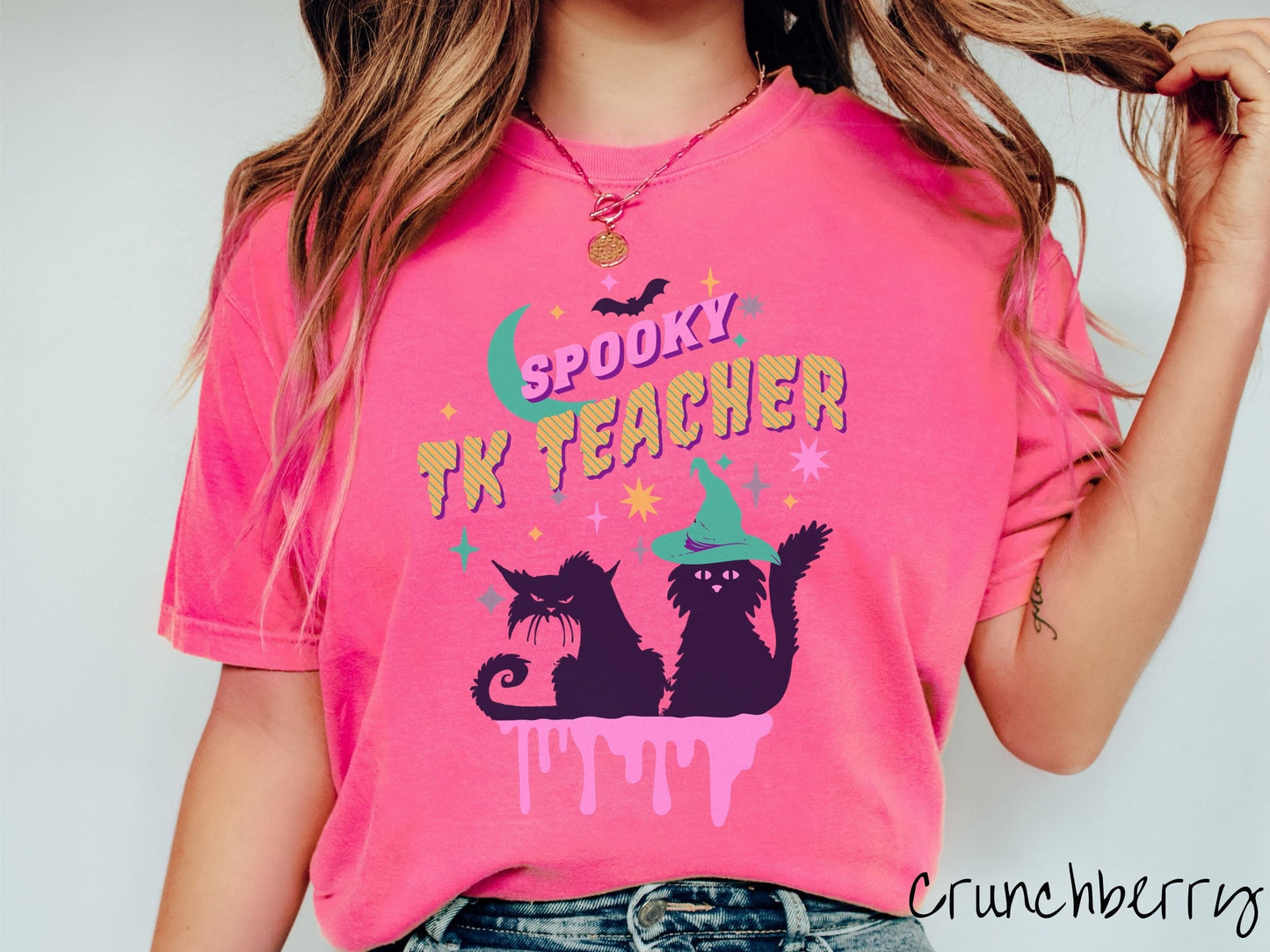 A woman wearing a cute, vintage crunchberry colored Comfort Colors t-shirt with the text Spooky TK Teacher in pink and yellow font. Below are two black cats with pink eyes wearing witch hats sitting beneath colorful stars and a bright green moon.