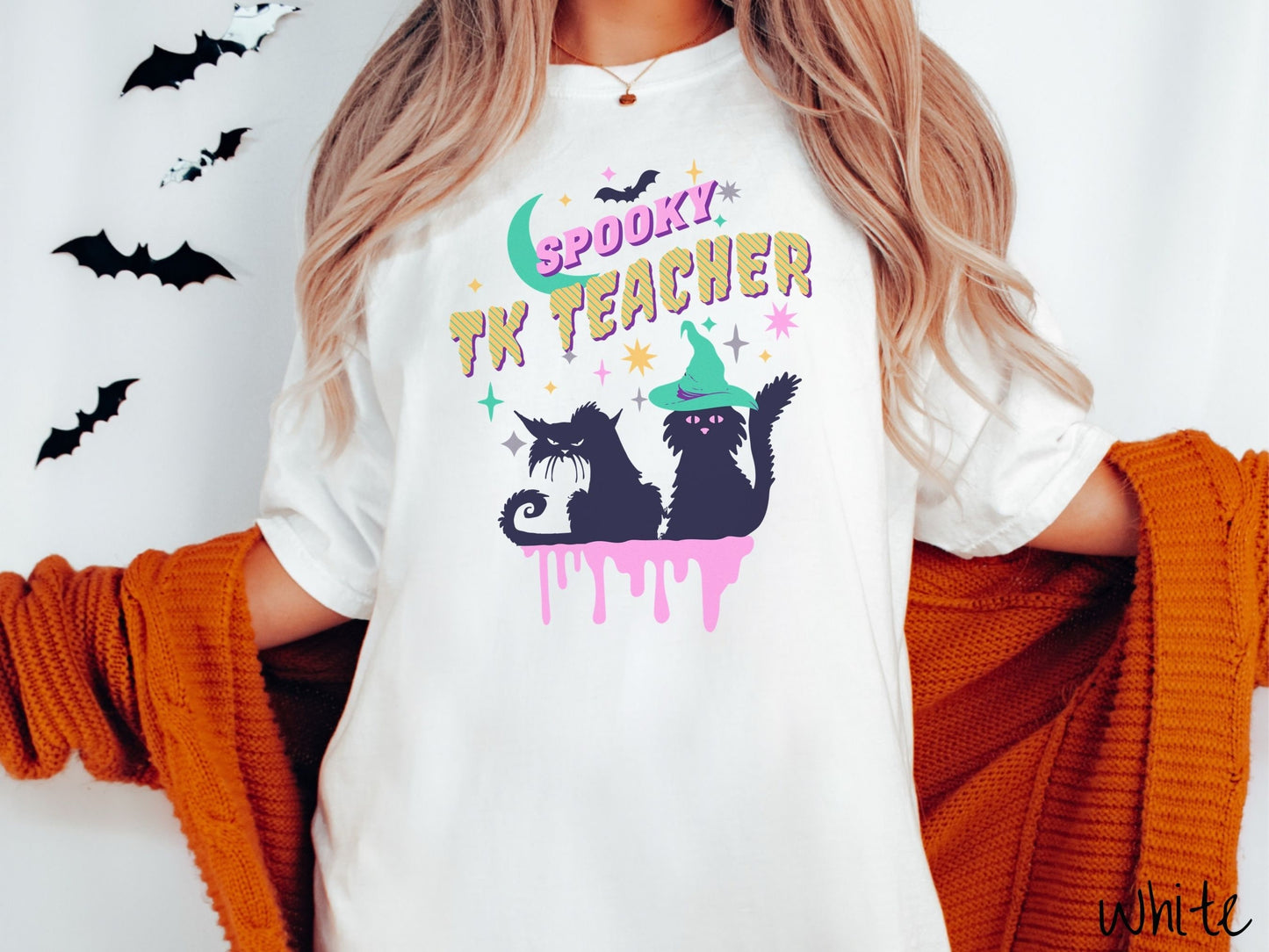 A woman wearing a cute, vintage white colored Comfort Colors t-shirt with the text Spooky TK Teacher in pink and yellow font. Below are two black cats with pink eyes wearing witch hats sitting beneath colorful stars and a bright green moon.