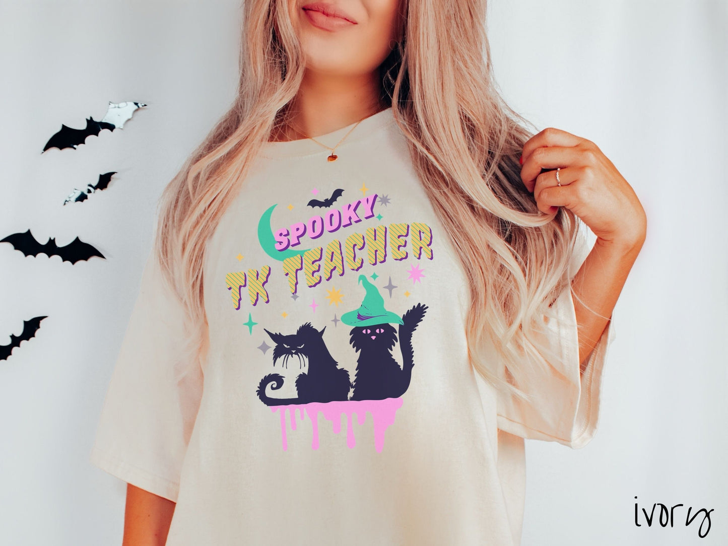 A woman wearing a cute, vintage ivory colored Comfort Colors t-shirt with the text Spooky TK Teacher in pink and yellow font. Below are two black cats with pink eyes wearing witch hats sitting beneath colorful stars and a bright green moon.