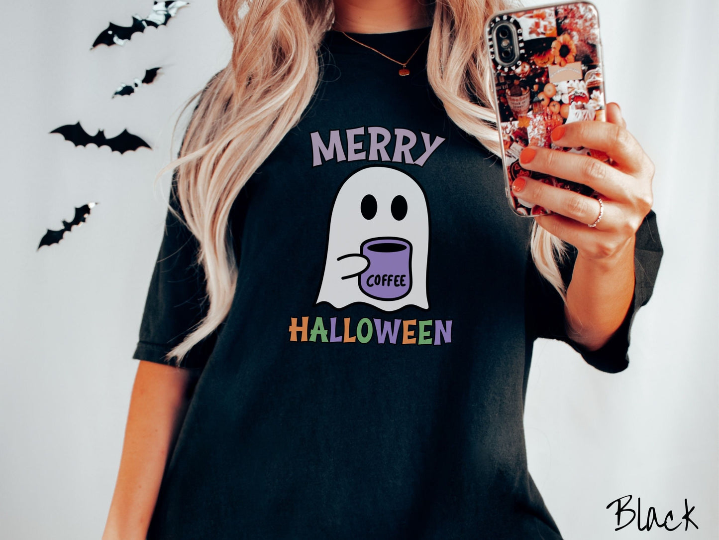 A woman wearing a cute, vintage black colored Comfort Colors t-shirt with the text Merry Halloween in purple, orange, and green font. There is a cute ghost holding a coffee mug in between the text.