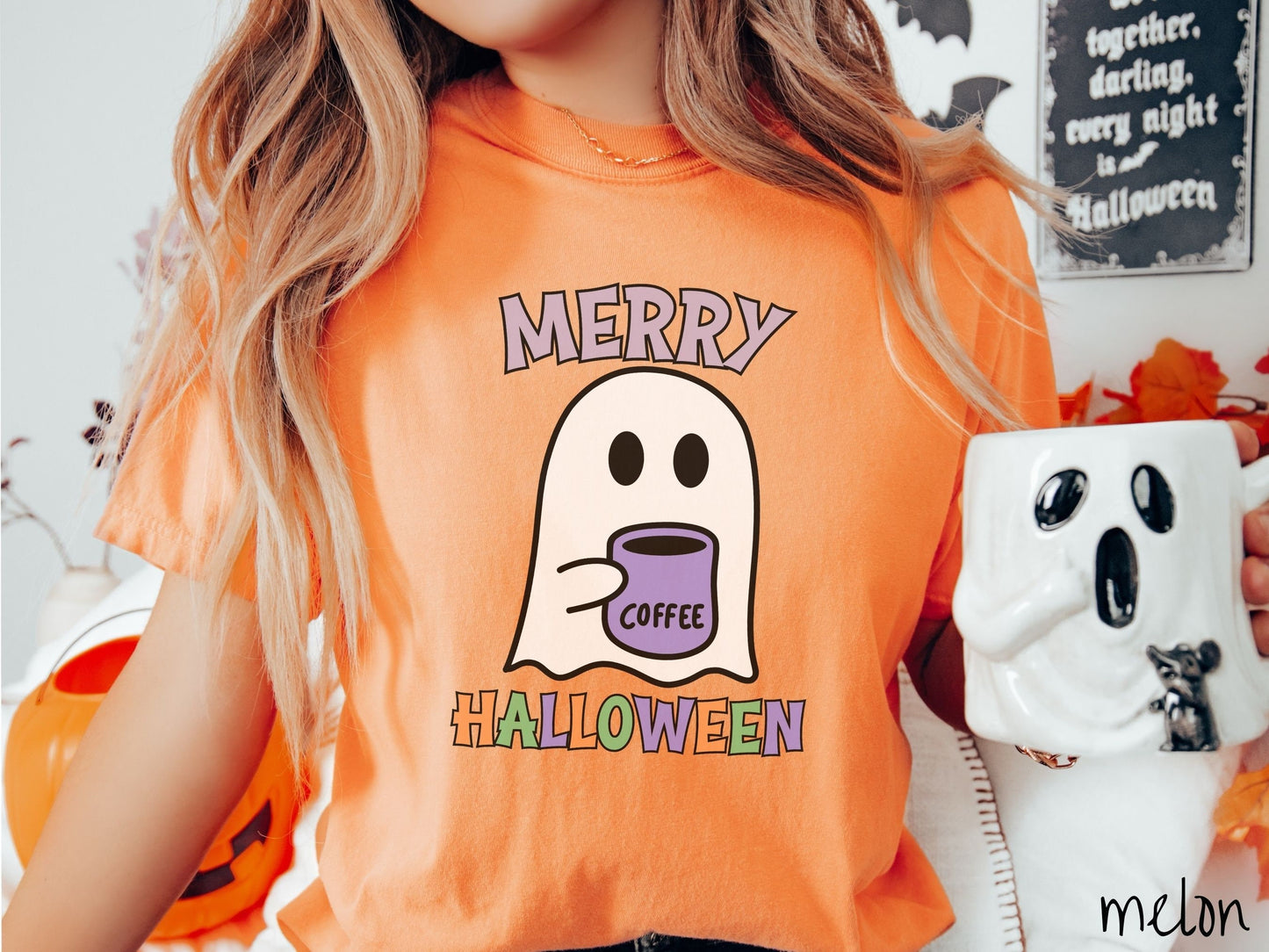 A woman wearing a cute, vintage melon colored Comfort Colors t-shirt with the text Merry Halloween in purple, orange, and green font. There is a cute ghost holding a coffee mug in between the text.