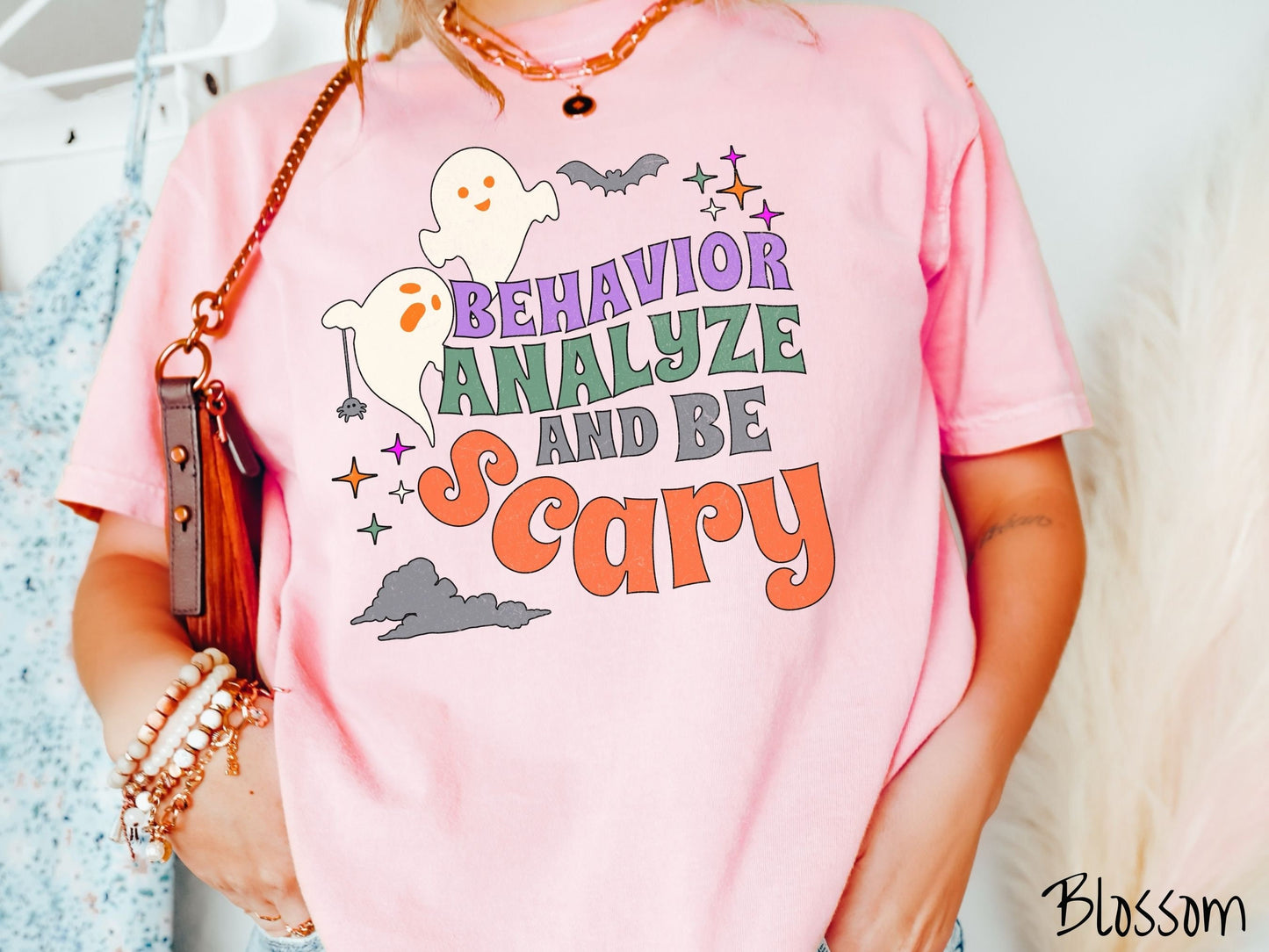A woman wearing a cute, vintage blossom colored Comfort Colors T-shirt with the text Behavior Analyze and Be Scary in colorful font. There are orange-eyed ghosts flying above and a black bat. A black spider is dangling by its web from one ghost.
