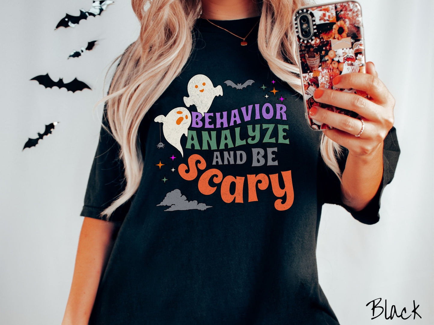 A woman wearing a cute, vintage black colored Comfort Colors T-shirt with the text Behavior Analyze and Be Scary in colorful font. There are orange-eyed ghosts flying above and a black bat. A black spider is dangling by its web from one ghost.