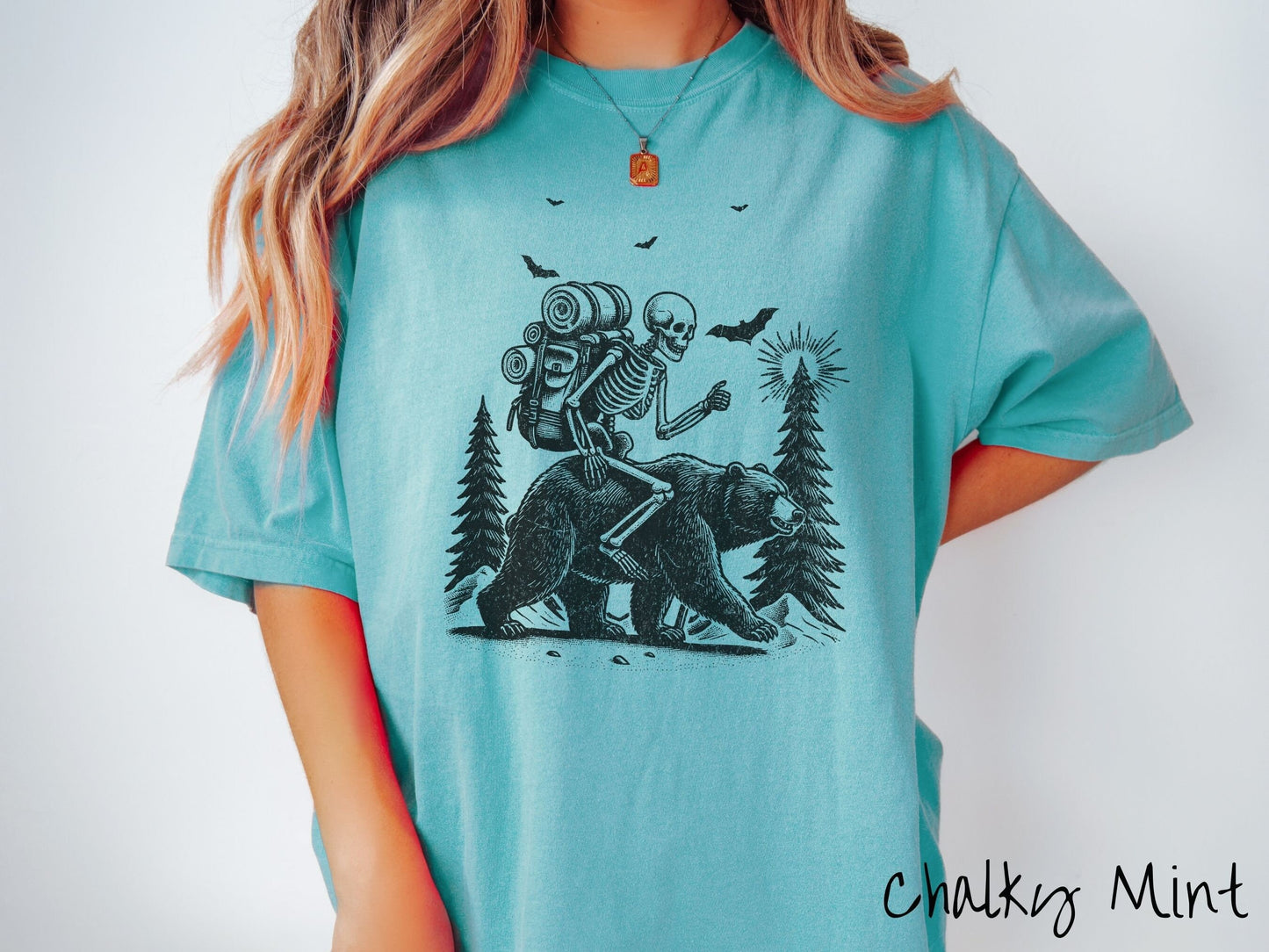 A woman wearing a cute, vintage chalky mint colored Comfort Colors T-shirt with a smiling skeleton wearing a camping backpack and riding a bear in the woods. There are trees in the background in front of the moon and black bats in the air.