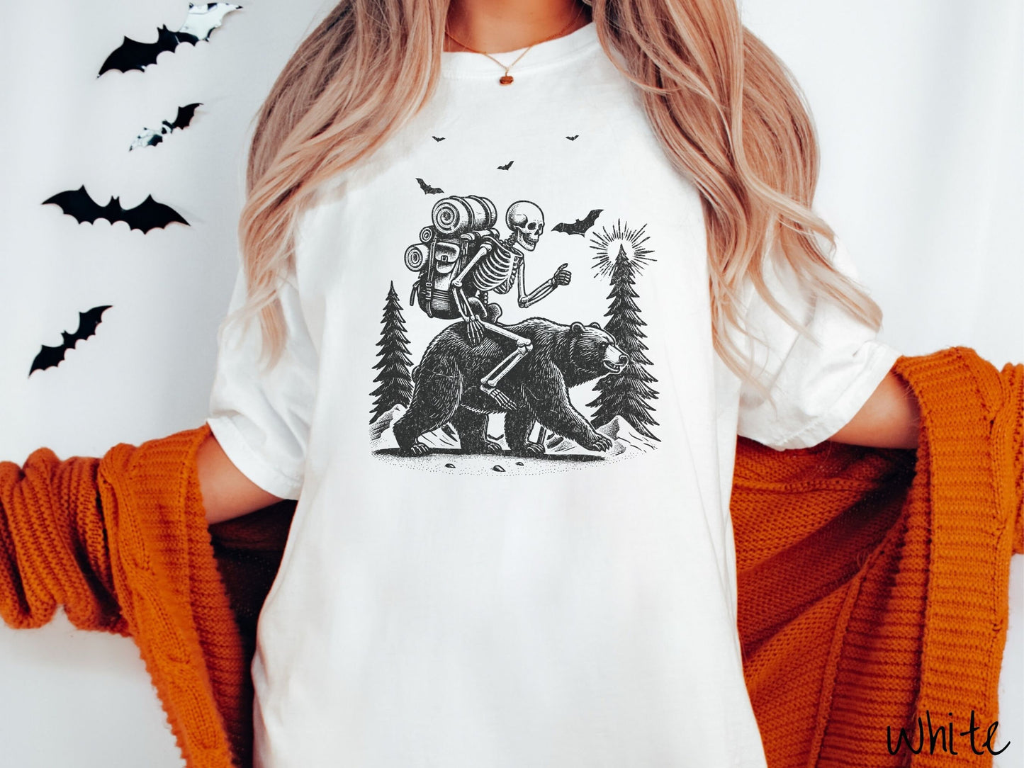 A woman wearing a cute, vintage white colored Comfort Colors T-shirt with a smiling skeleton wearing a camping backpack and riding a bear in the woods. There are trees in the background in front of the moon and black bats in the air.