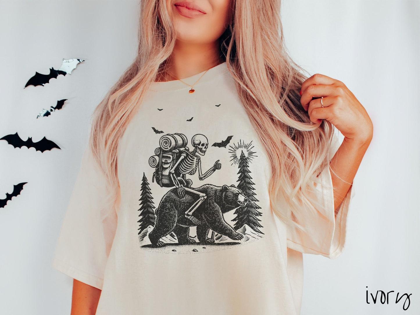 A woman wearing a cute, vintage ivory colored Comfort Colors T-shirt with a smiling skeleton wearing a camping backpack and riding a bear in the woods. There are trees in the background in front of the moon and black bats in the air.