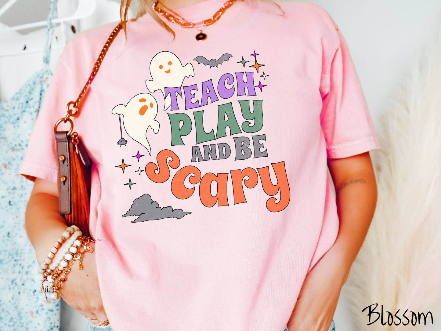 A woman wearing a cute, vintage blossom colored Comfort Colors T-shirt with the text Teach Play and Be Scary in colorful font. There are orange-eyed white ghosts flying around and black bats and twinkling stars in the sky.