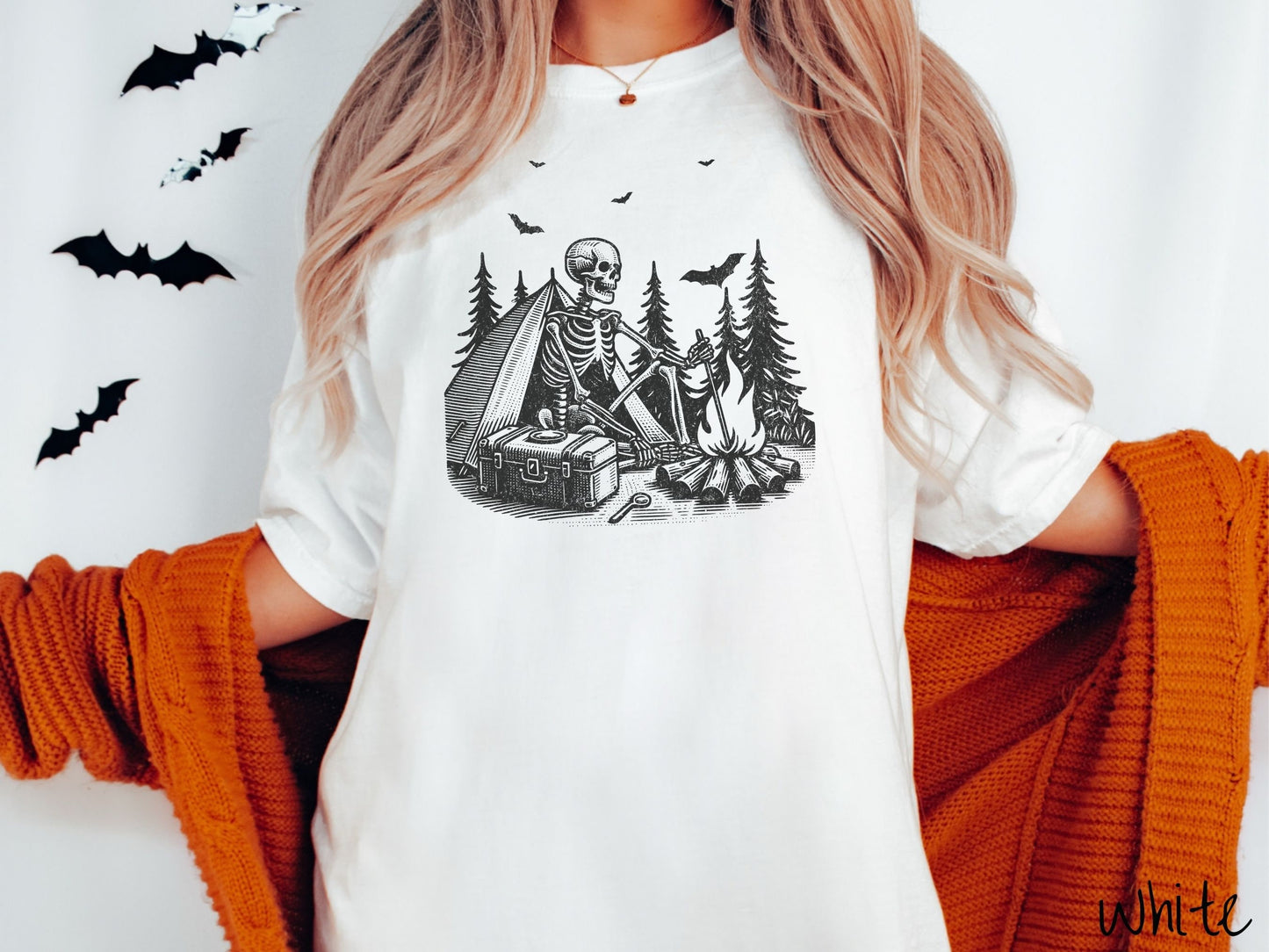 A woman wearing a cute, vintage white colored Comfort Colors T-shirt with a skeleton sitting in a tent building a fire next to a cooler out in the woods. The campsite is surrounded by evergreen trees and there are bats flying above.