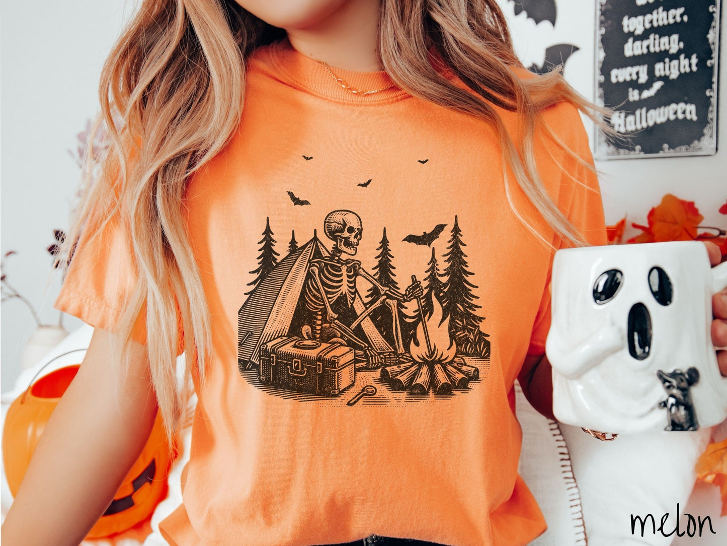 A woman wearing a cute, vintage melon colored Comfort Colors T-shirt with a skeleton sitting in a tent building a fire next to a cooler out in the woods. The campsite is surrounded by evergreen trees and there are bats flying above.