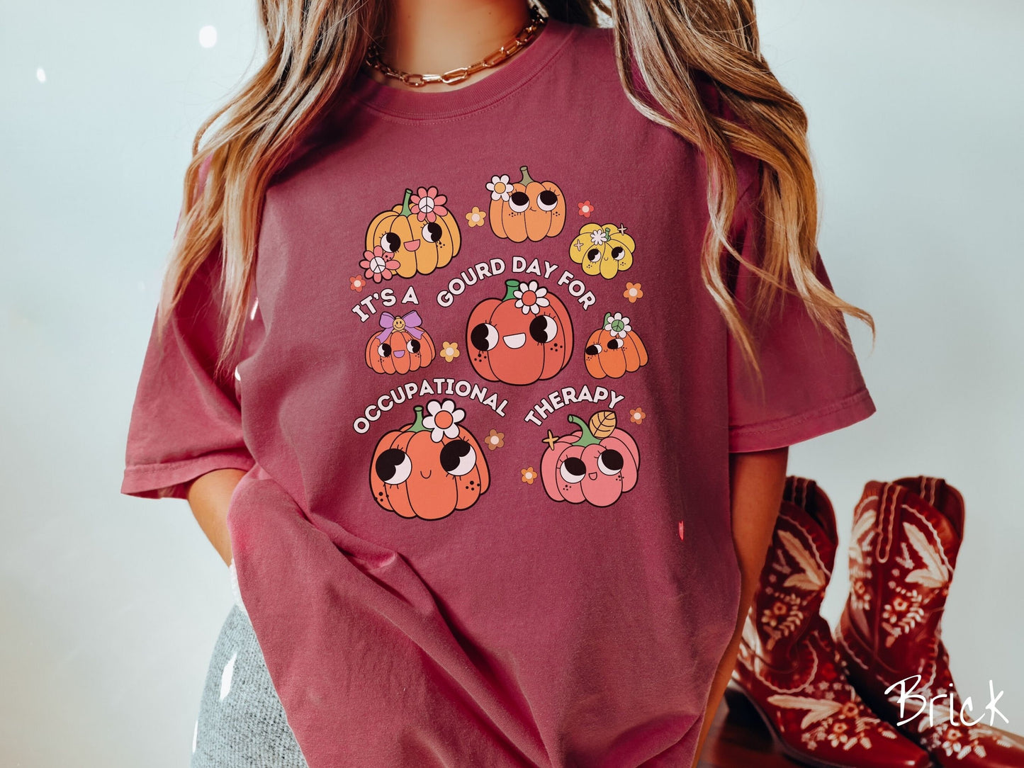 A woman wearing a cute, vintage brick colored Comfort Colors T-shirt with text Its a Gourd Day for Occupational Therapy in white font. There is a three by three grid of orange, yellow, and pink pumpkins with girly eyes, wearing flowers and bows.