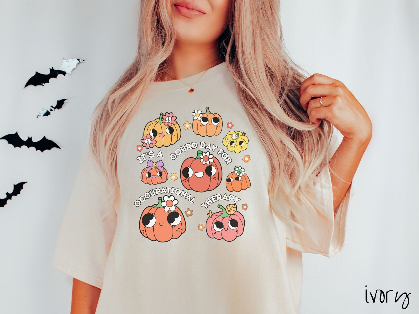 A woman wearing a cute, vintage ivory colored Comfort Colors T-shirt with text Its a Gourd Day for Occupational Therapy in white font. There is a three by three grid of orange, yellow, and pink pumpkins with girly eyes, wearing flowers and bows.