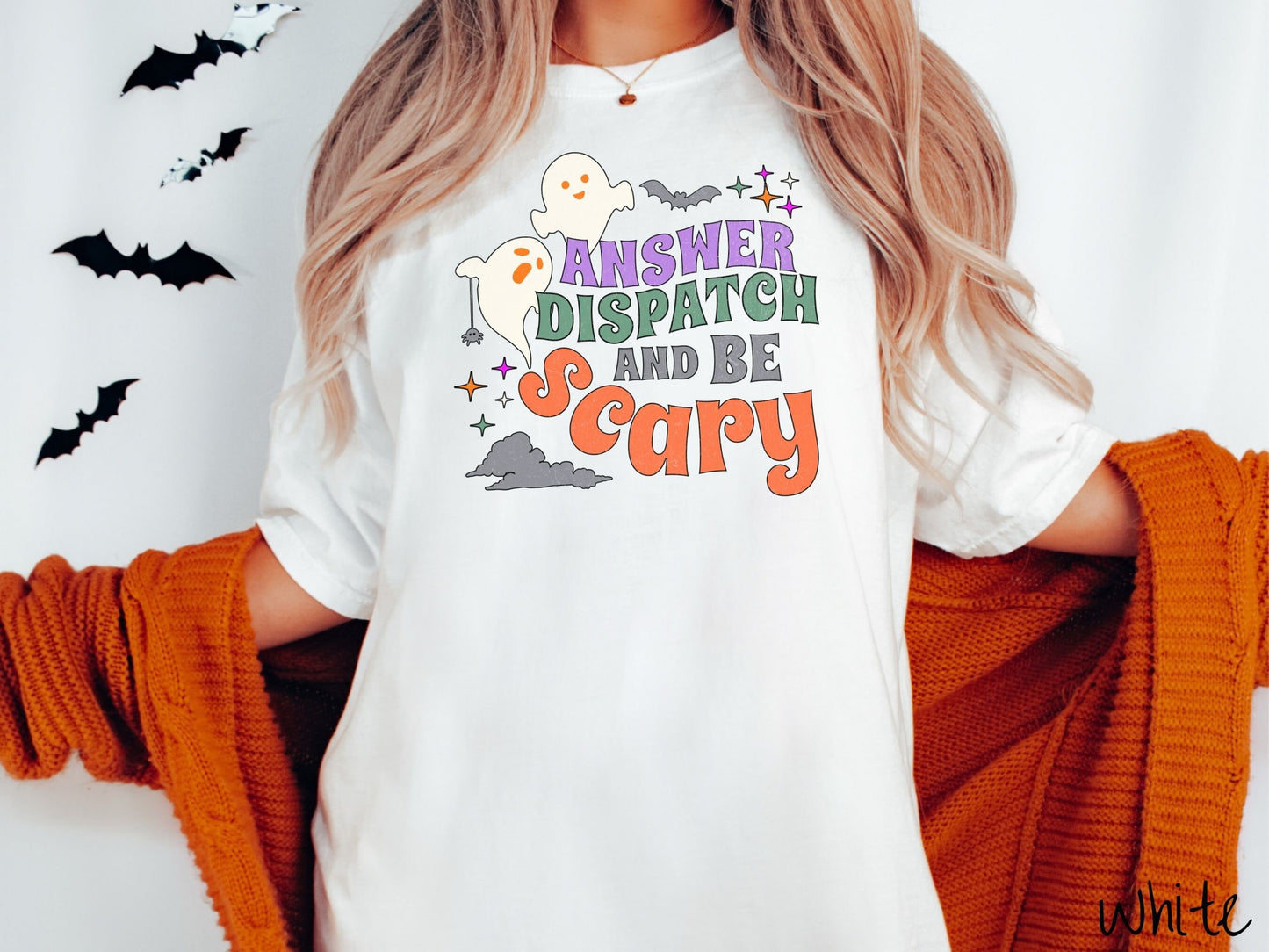 A woman wearing a cute, vintage white colored Comfort Colors T-shirt with the text Answer Dispatch and Be scary in colorful font. There are orange-eyed white ghosts flying around and black bats and twinkling stars in the sky.