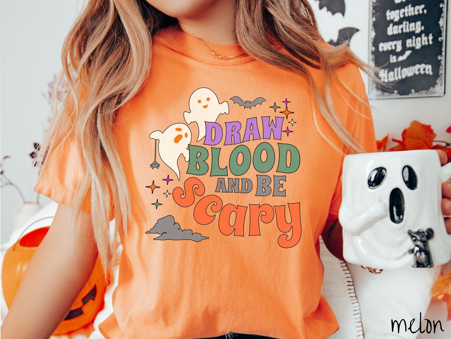 A woman wearing a cute, vintage melon colored Comfort Colors T-shirt with the text Draw Blood and Be Scary in colorful font. There are orange-eyed white ghosts flying around and black bats and twinkling stars in the sky.