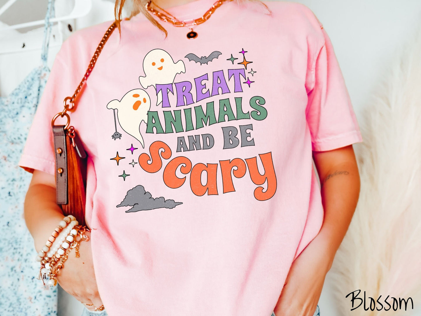 A woman wearing a cute, vintage blossom colored Comfort Colors T-shirt with the text Treat Animals and Be Scary in colorful font. There are orange-eyed white ghosts flying around and black bats and twinkling stars in the sky.