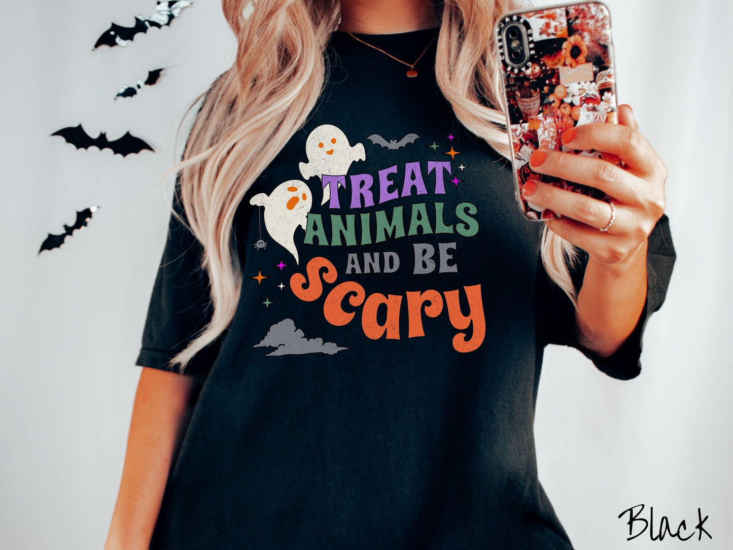A woman wearing a cute, vintage black colored Comfort Colors T-shirt with the text Treat Animals and Be Scary in colorful font. There are orange-eyed white ghosts flying around and black bats and twinkling stars in the sky.