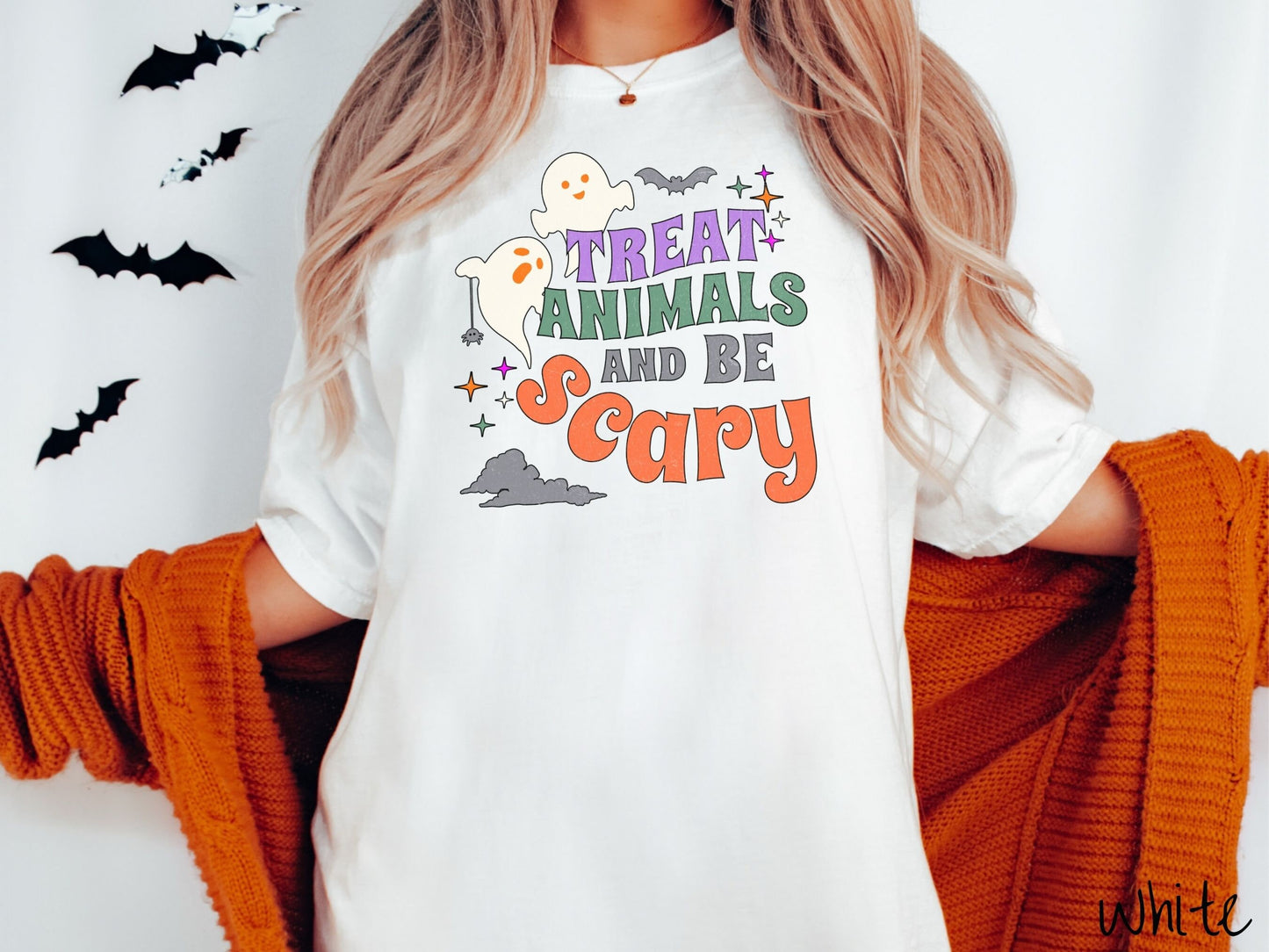 A woman wearing a cute, vintage white colored Comfort Colors T-shirt with the text Treat Animals and Be Scary in colorful font. There are orange-eyed white ghosts flying around and black bats and twinkling stars in the sky.
