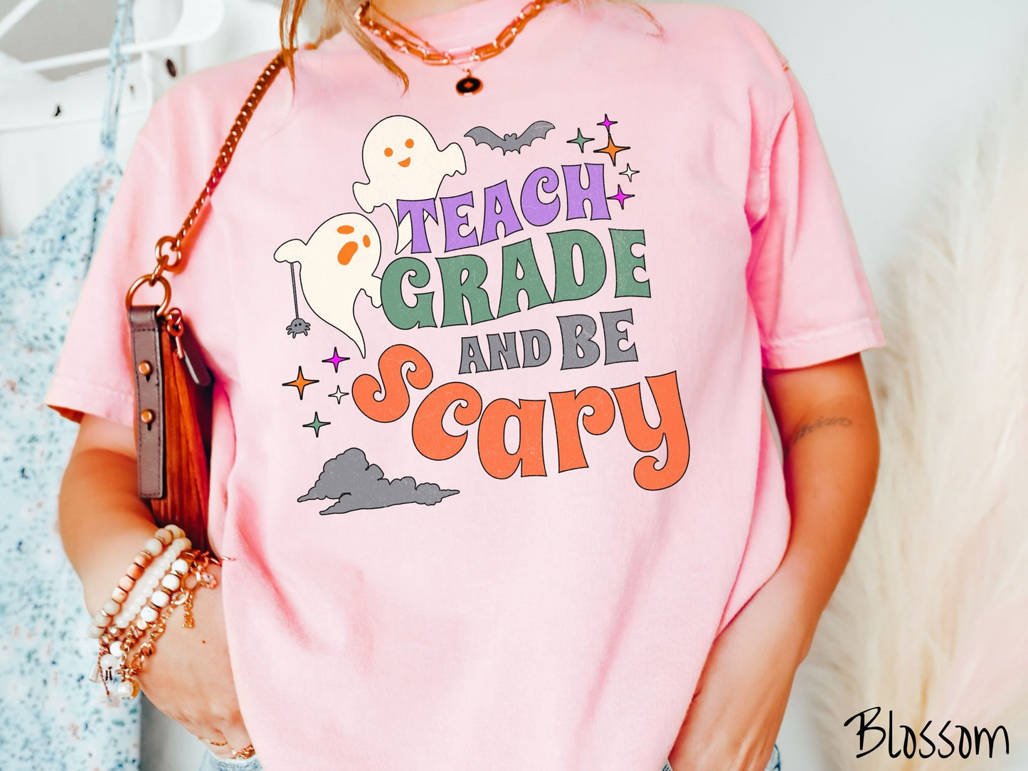 A woman wearing a cute, vintage blossom colored Comfort Colors T-shirt with the text Teach Grade and Be Scary in colorful font. There are orange-eyed white ghosts flying around and black bats and twinkling stars in the sky.