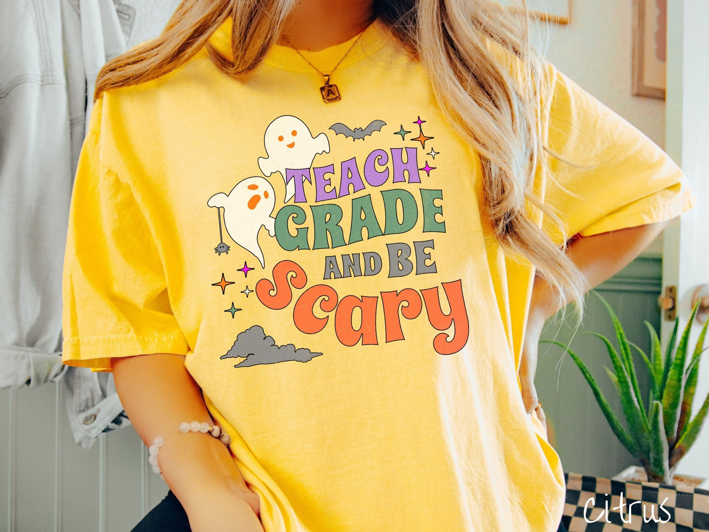 A woman wearing a cute, vintage citrus colored Comfort Colors T-shirt with the text Teach Grade and Be Scary in colorful font. There are orange-eyed white ghosts flying around and black bats and twinkling stars in the sky.